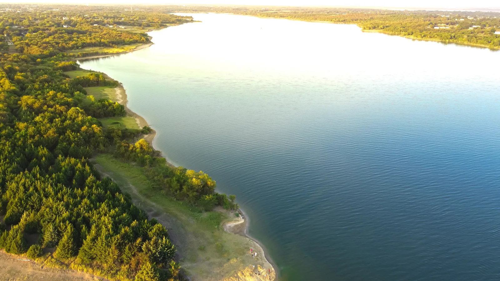 Aerial view of a wide, calm lake bordered by a dense green forest under bright sunlight.