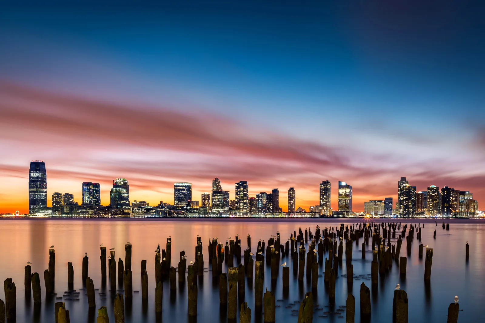 A wide view of an illuminated urban skyline across calm water at sunset, with wooden pier pilings standing in the foreground.