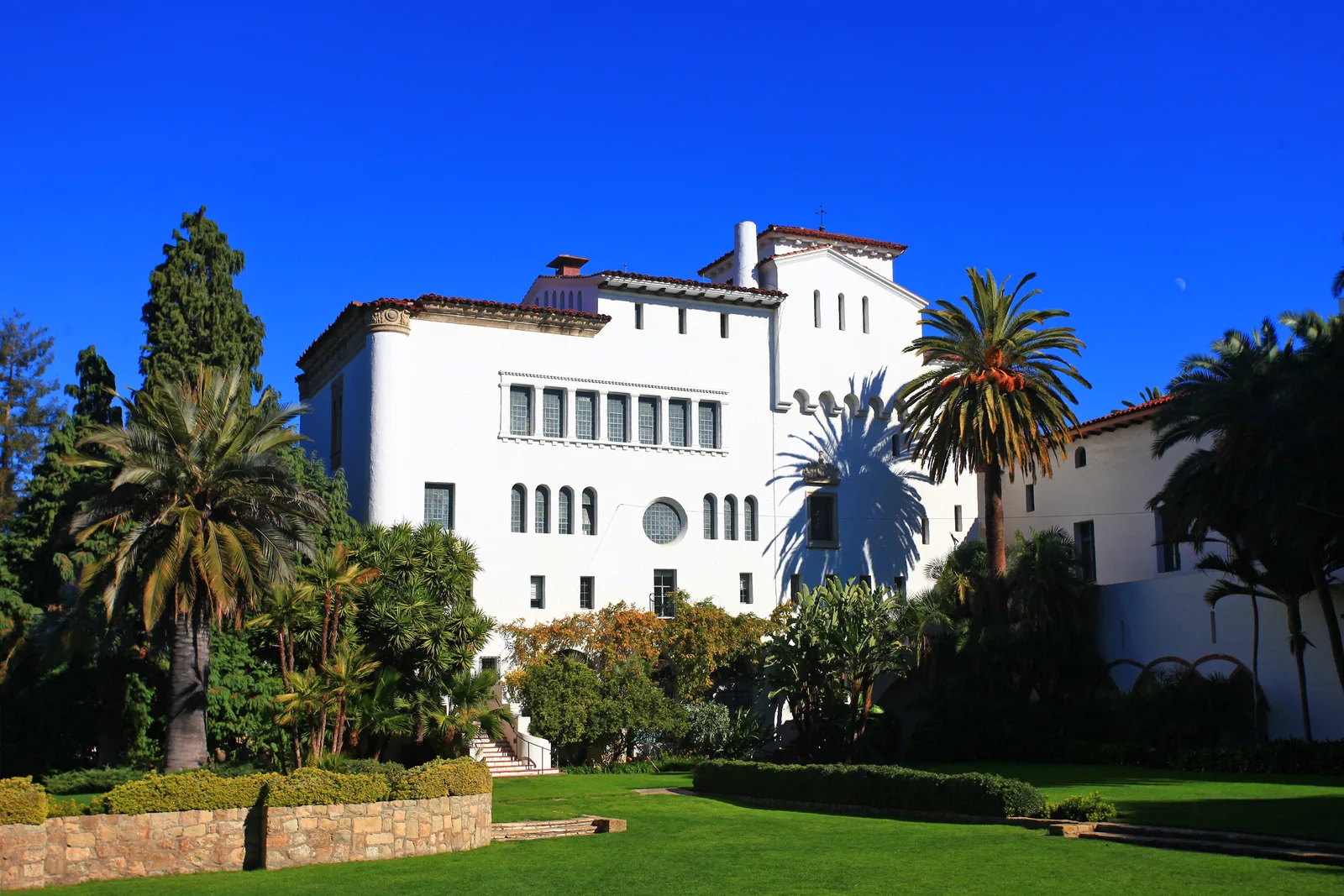 The historic white Spanish-Moorish Revival style Santa Barbara County Courthouse stands surrounded by lush green lawns and towering palm trees under a bright blue sky