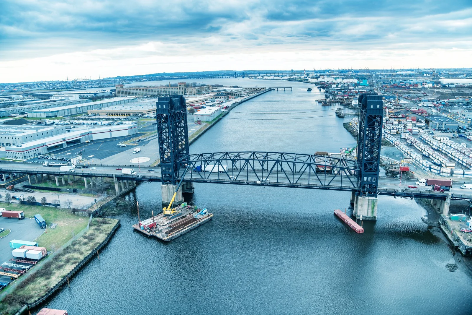 Aerial view of a large steel vertical-lift bridge spanning an industrial riverway with construction barges positioned at its base.