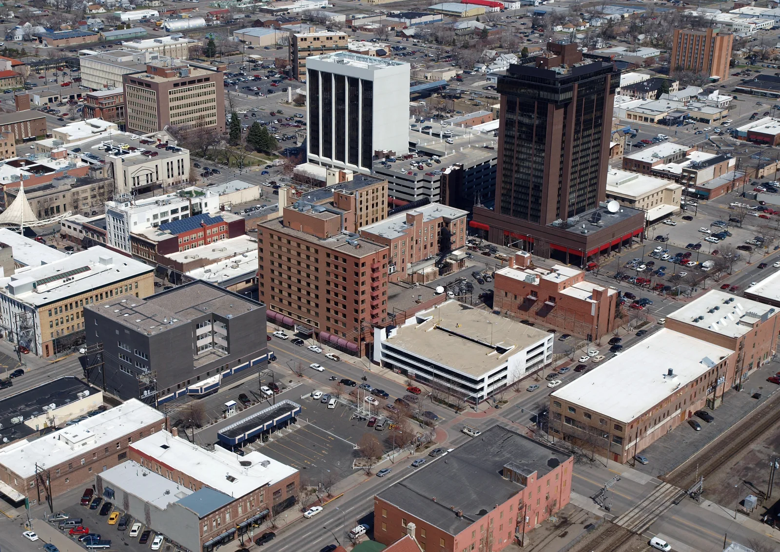 Aerial view of a city center with various buildings, streets, and parking lots. A dark high-rise building and a white mid-rise building are prominent among other commercial and residential structures.