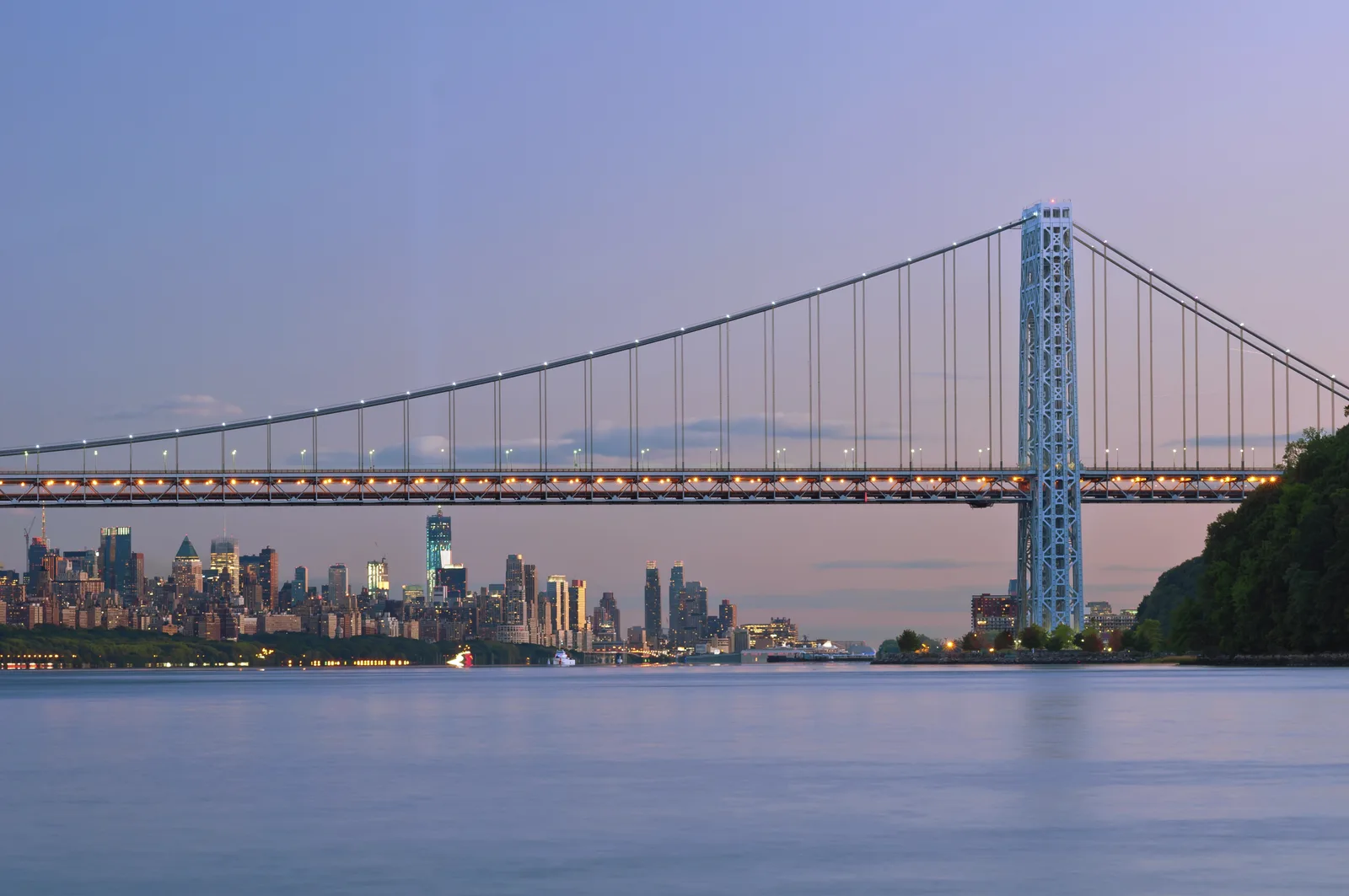Illuminated suspension bridge over a river at dusk with a city skyline in the background.