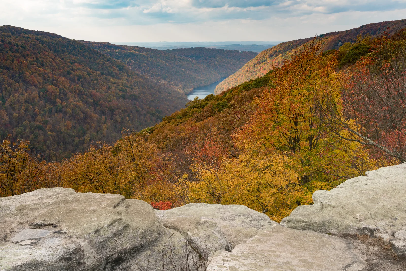 A rocky cliff overlook provides a scenic view of a river winding through a forested valley displaying vibrant autumn foliage.