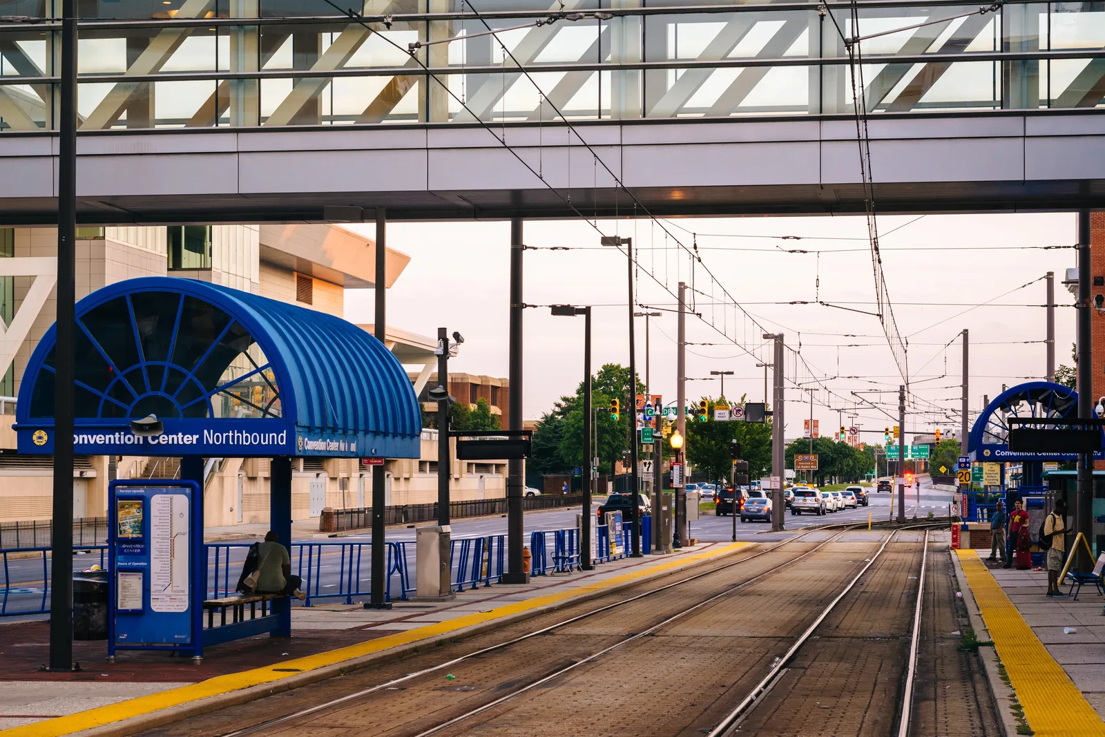 A light rail transit station platform featuring tracks, overhead wires, and blue passenger shelters.