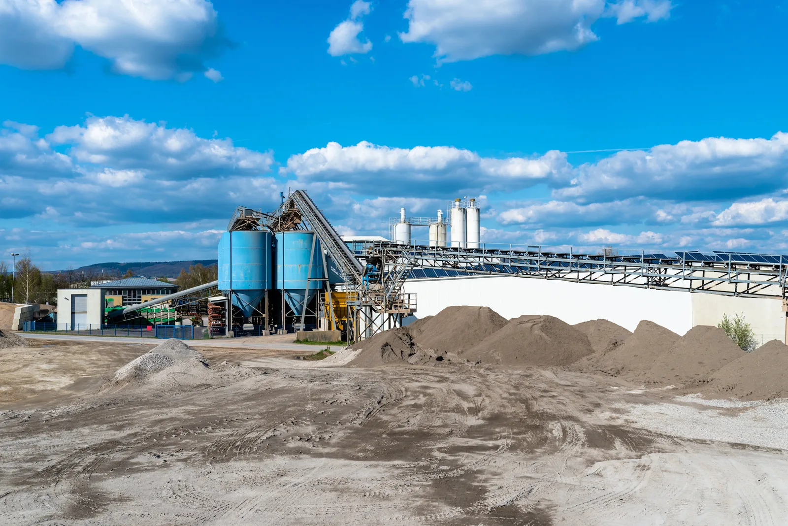 Industrial processing plant with blue storage silos, an inclined conveyor, and material stockpiles.