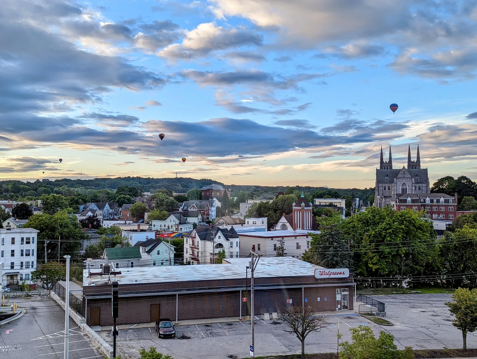 An aerial view of the Lewiston, Maine skyline featuring the historic Basilica of Saints Peter and Paul and hot air balloons under a partly cloudy sky.